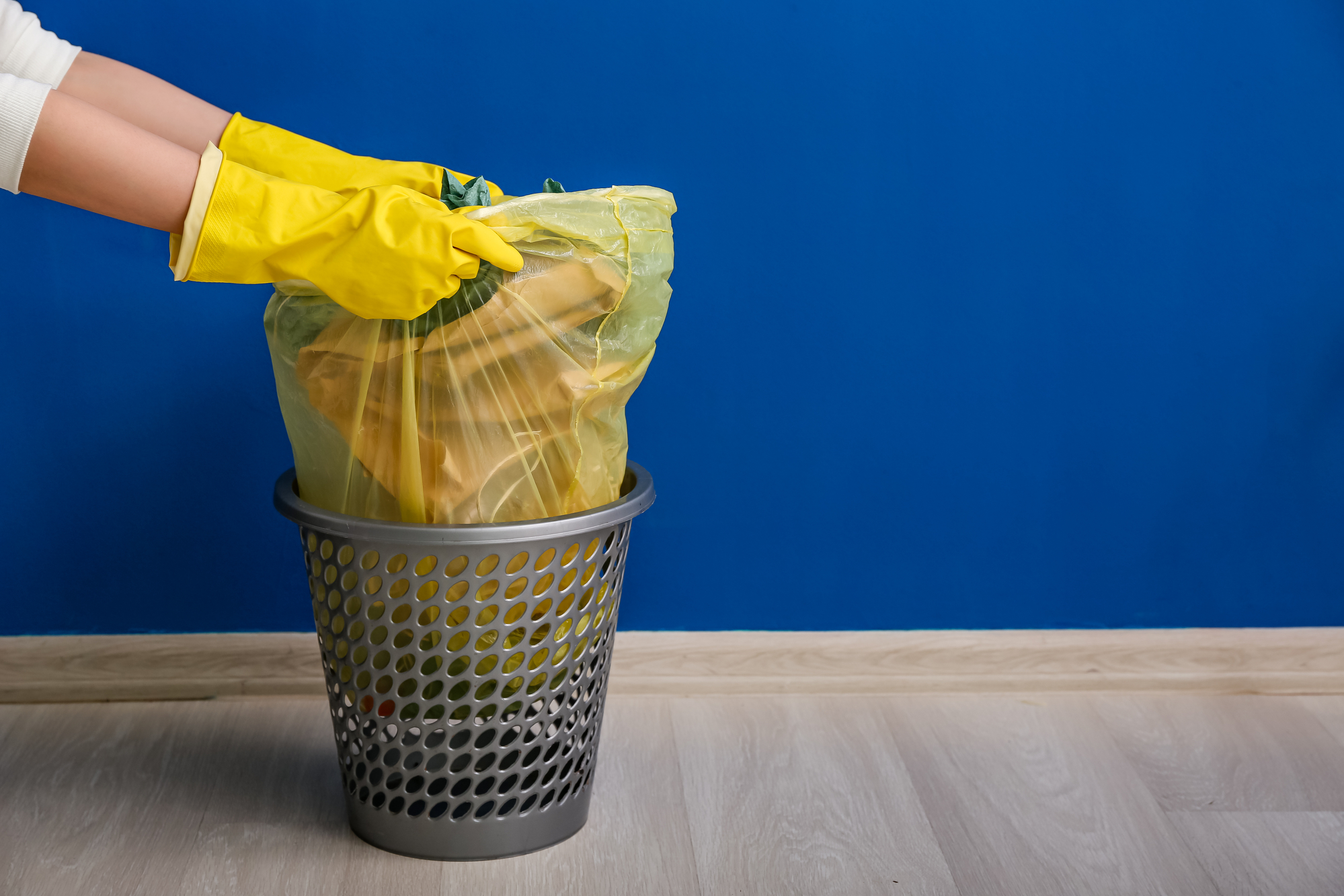 Female hands in rubber gloves pulling full garbage bag from trash bin near blue wall