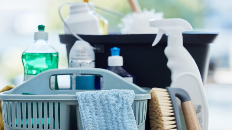 Basket, table and product for cleaning home with brush, cloth and chemical for hygiene by blurred background. Empty house, desk and plastic container for services, stop bacteria and dust in interior.