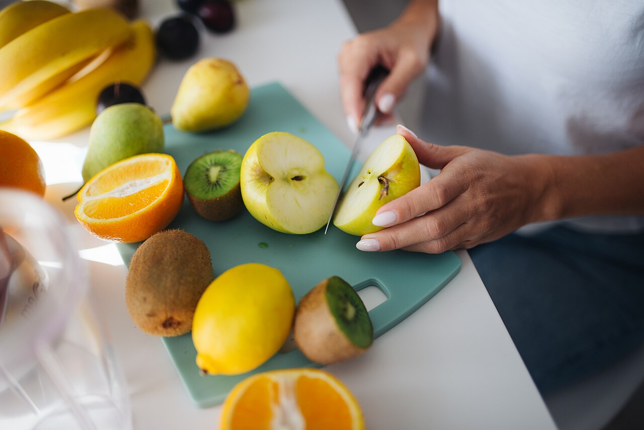 A person is cutting a green apple on a cutting board surrounded by various fresh fruits like lemons, oranges, and kiwis. The kitchen is well-lit and inviting.