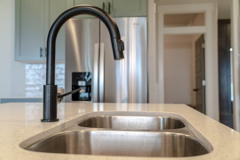 Double basin undermount kitchen sink with black faucet against refrigerator. Wall cabinets and open door can also be seen from the deep stainless steel island sink.