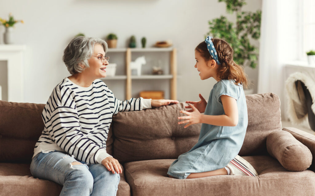 Little girl smiling and talking with astonished elderly woman while sitting on sofa at home together grandma babysitting